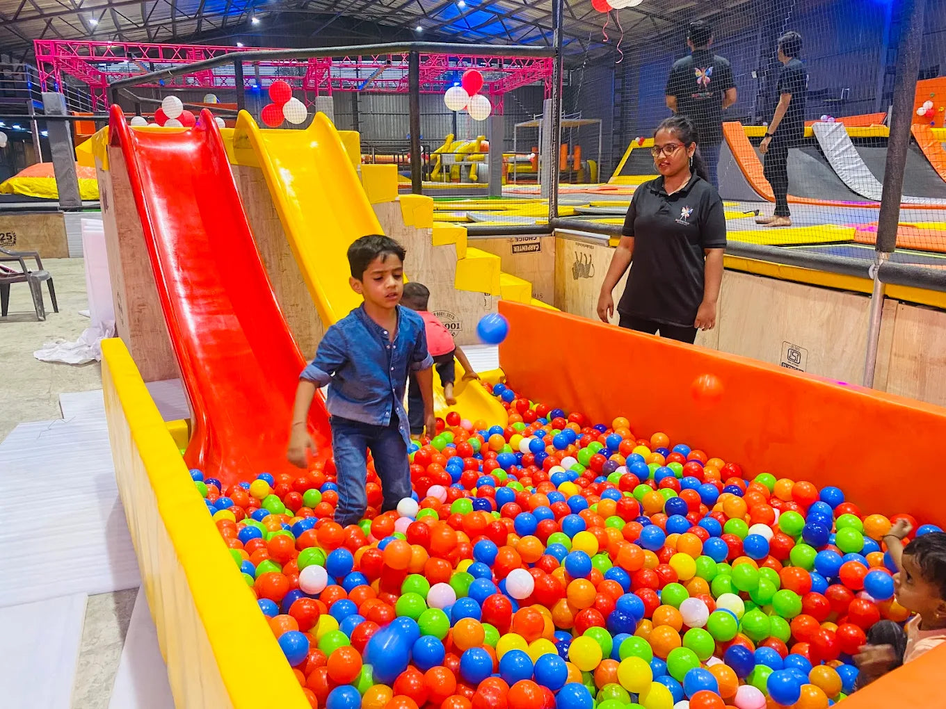 Children play in a ball pit at an indoor trampoline park with slides.