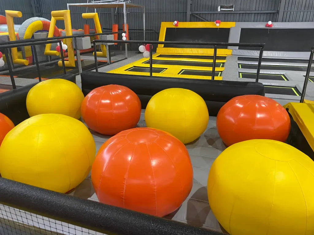 Large inflatable balls in yellow and orange are scattered on the floor of an indoor trampoline park.