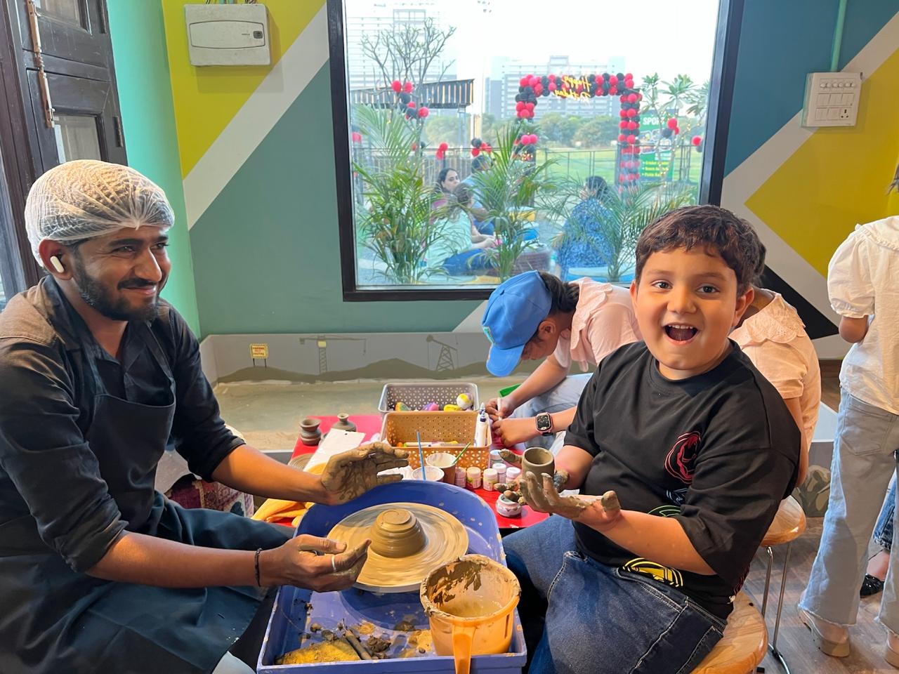 A man with clay on his hands guides a child at a pottery wheel, while other children are visible in the background.