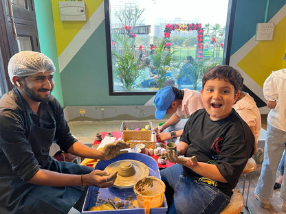 A man with clay on his hands guides a child at a pottery wheel, while other children are visible in the background.