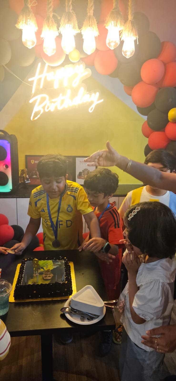 Children gather around a birthday cake with a picture of a soccer player, celebrating a birthday party.