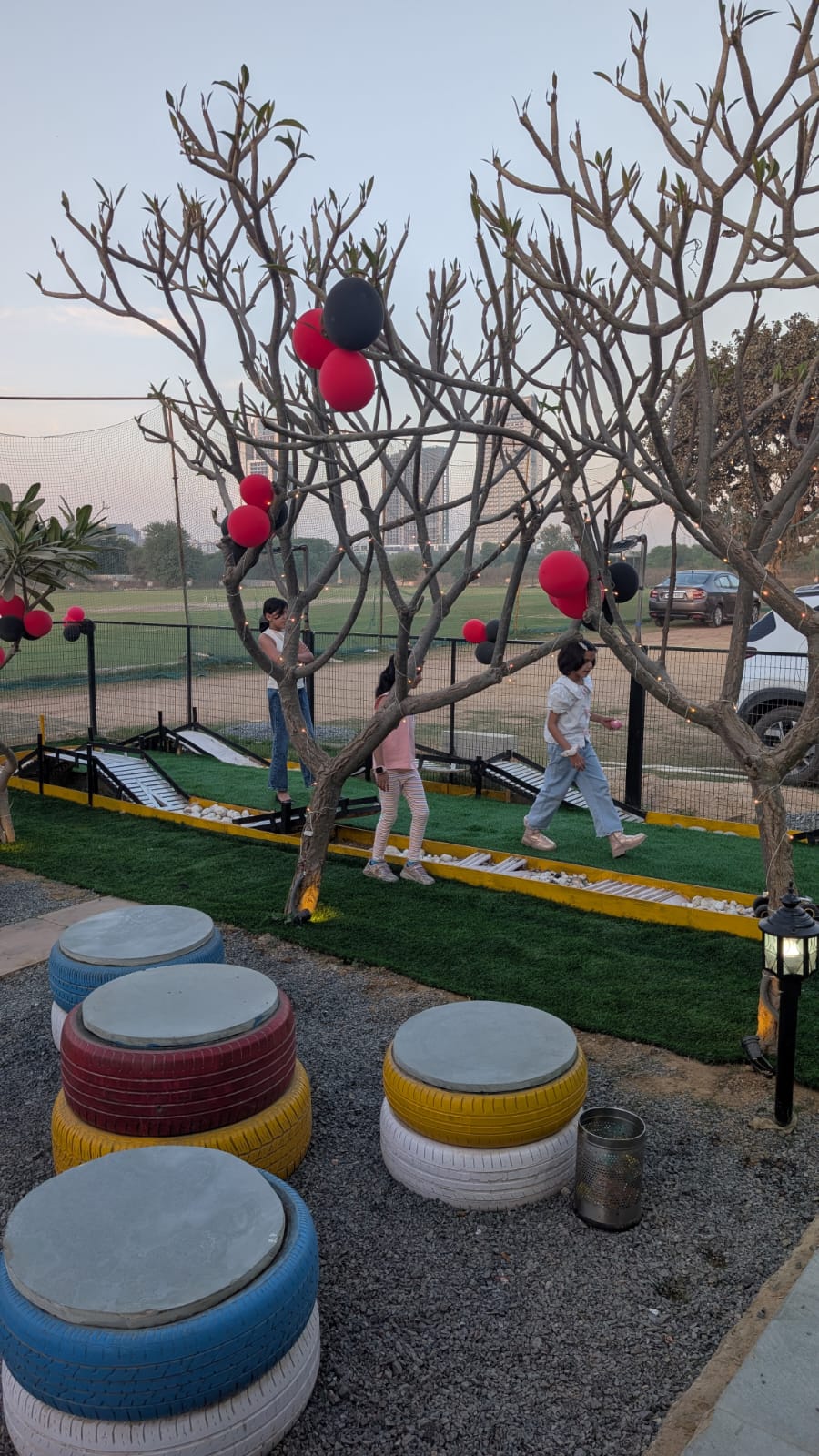 Children play mini-golf on a grassy course decorated with balloons and tire seating.