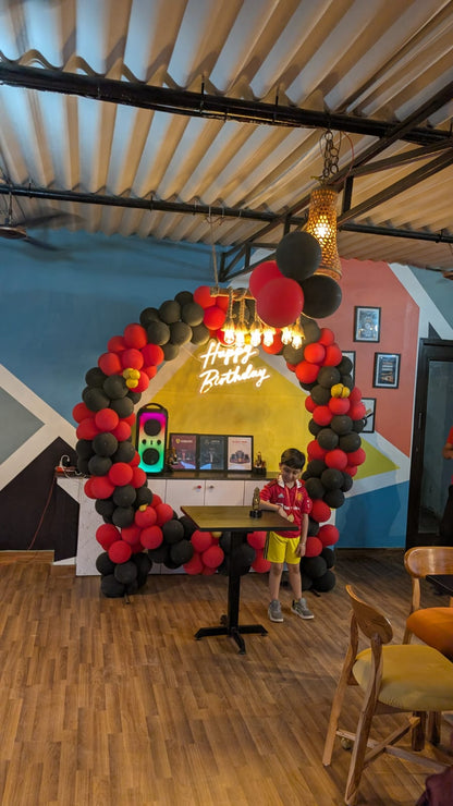 A young boy in a soccer jersey stands by a table at a birthday party decorated with a balloon arch and "Happy Birthday" neon sign.