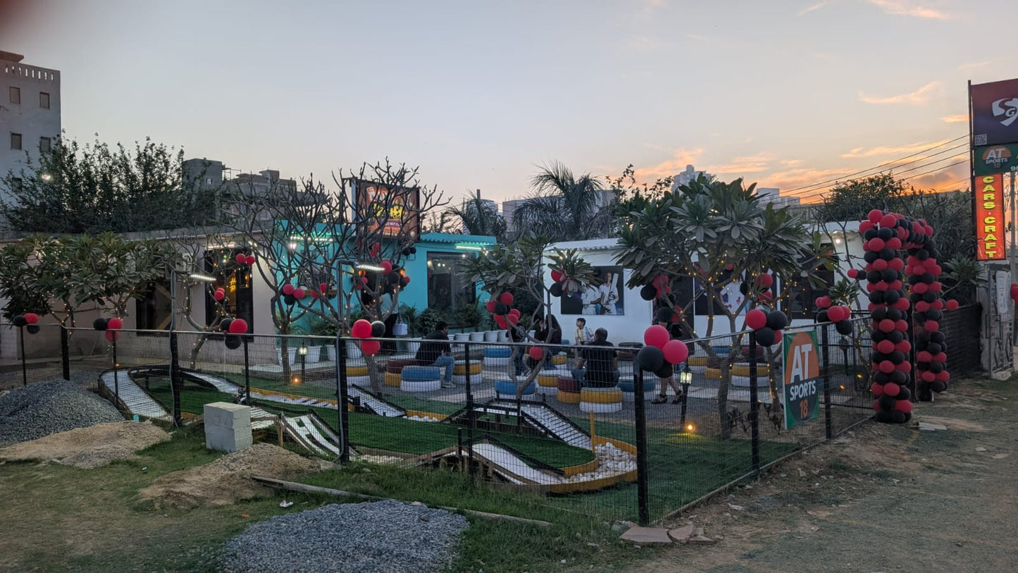 An outdoor mini-golf course with people of various ages enjoying the game, decorated with red and black balloons.
