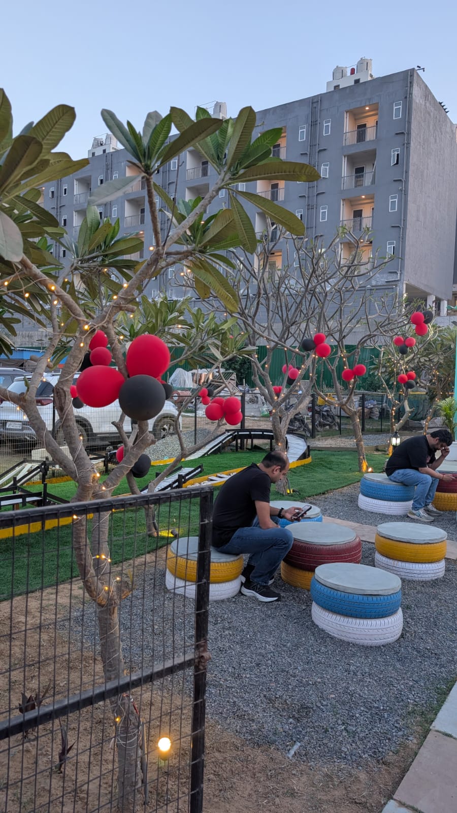 Two young men sit on colorful tire seats in an outdoor cafe with trees decorated with lights and balloons.