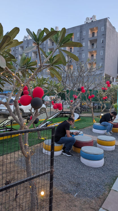 Two young men sit on colorful tire seats in an outdoor cafe with trees decorated with lights and balloons.