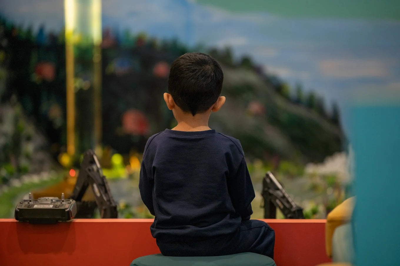 A young child sits facing away, looking at a miniature landscape with toy vehicles.