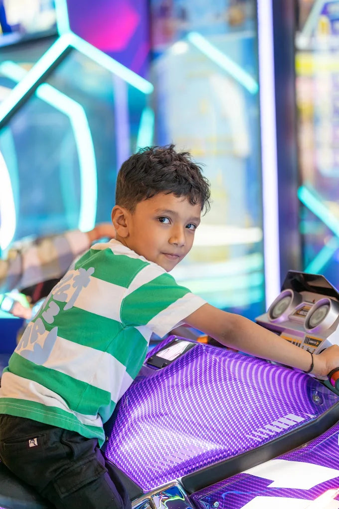 A young boy in a green and white striped shirt sits on a purple motorcycle arcade game.
