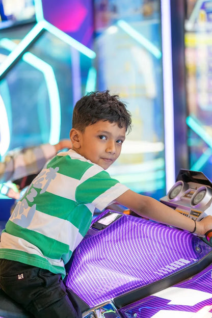 A young boy in a green and white striped shirt sits on a purple motorcycle arcade game.