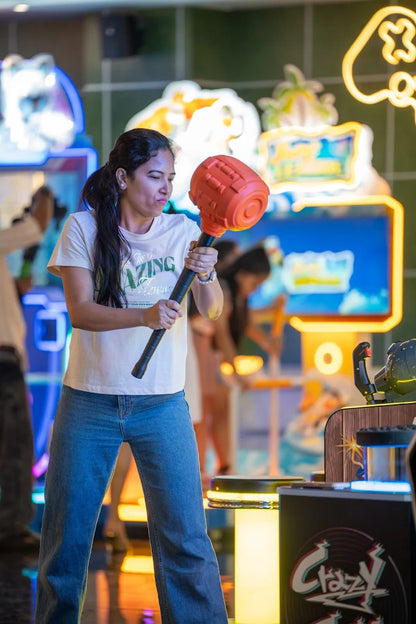 A woman plays an arcade game, swinging a large toy hammer. She is in an arcade with neon lights and other games.