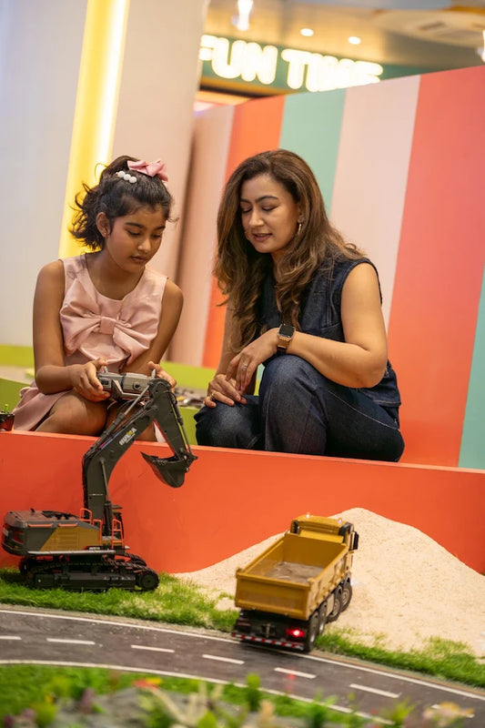 A young girl and a woman play with toy construction vehicles in a colorful indoor setting.