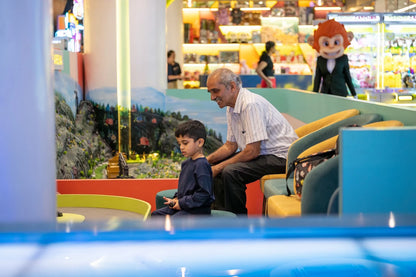 An older man and a young boy sit together in a brightly lit arcade, looking at a game. A mascot in a monkey costume stands in the background.