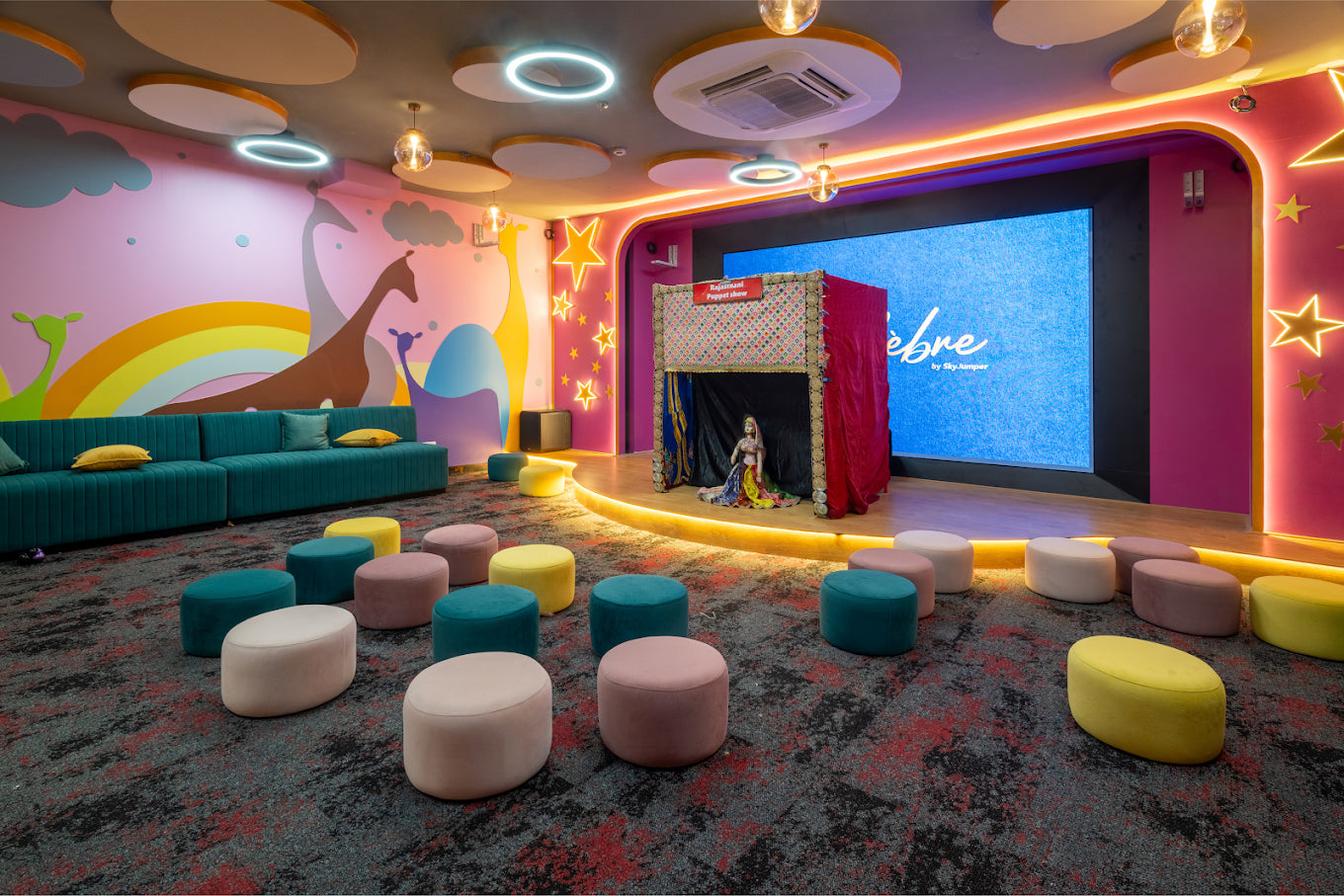 A colorful children's play area with a puppet show stage, plush seating, and a large screen.