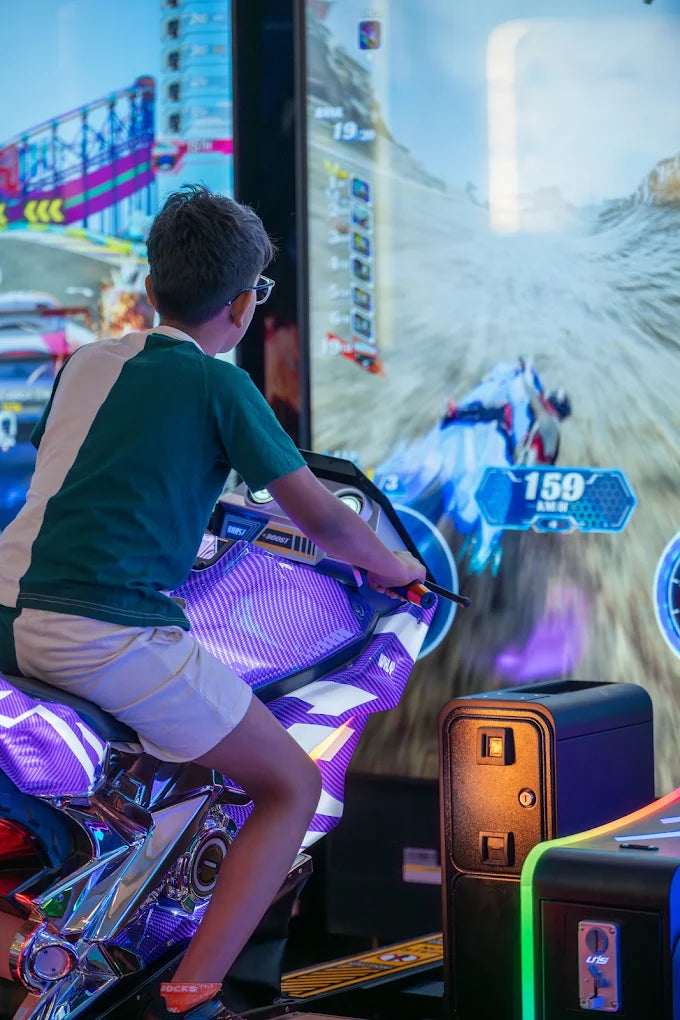A young boy plays a motorcycle racing arcade game, focused on the screen. The game features a vibrant, futuristic racing environment.