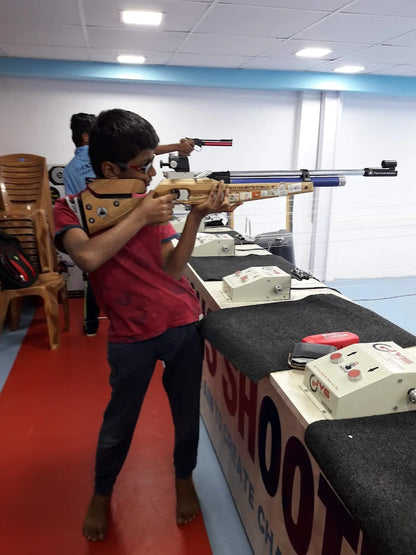 A young boy aims a rifle at a shooting range, with another person in the background also holding a firearm.