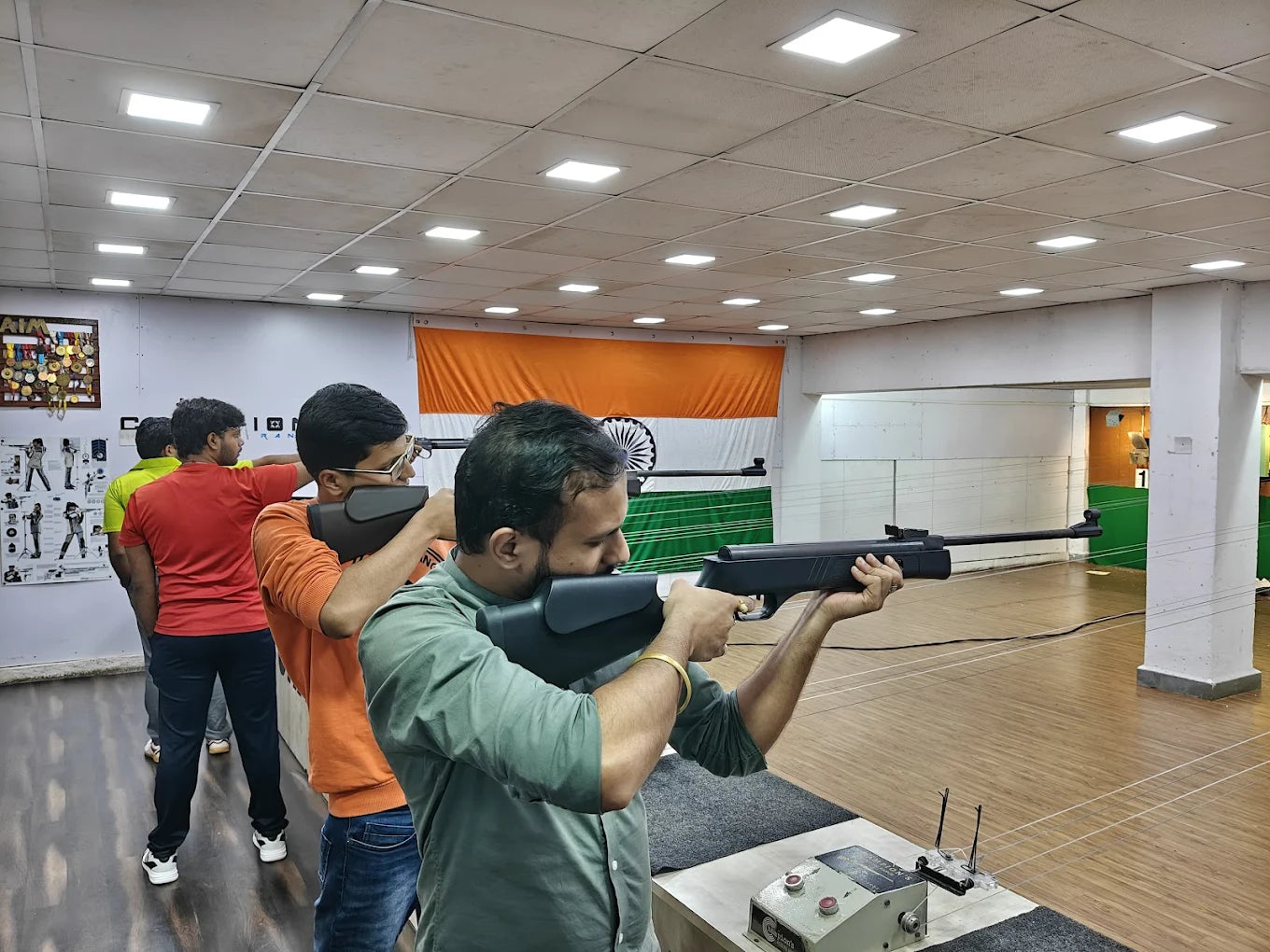 Young men practice shooting with air rifles in an indoor range, with an Indian flag displayed in the background.
