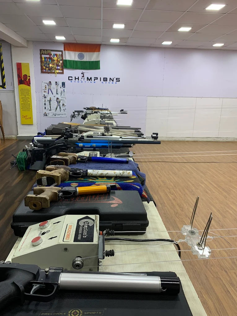 Rifles are lined up on tables at a shooting range, with an Indian flag and medals on the wall.
