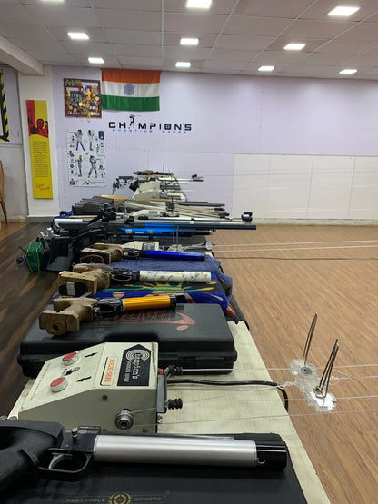 Rifles are lined up on tables at a shooting range, with an Indian flag and medals on the wall.
