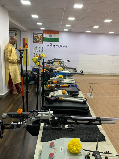 A man in traditional Indian attire stands by a row of competition air rifles at a shooting range.