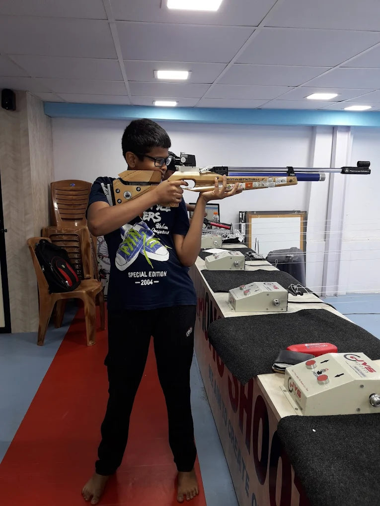 A young person aims a rifle at a shooting range. They are wearing glasses and standing barefoot on a red mat.