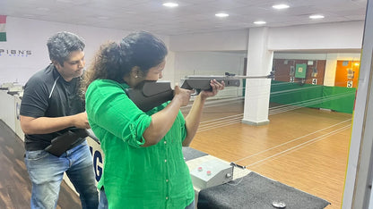 A woman practices shooting with a rifle at an indoor range, guided by a man.