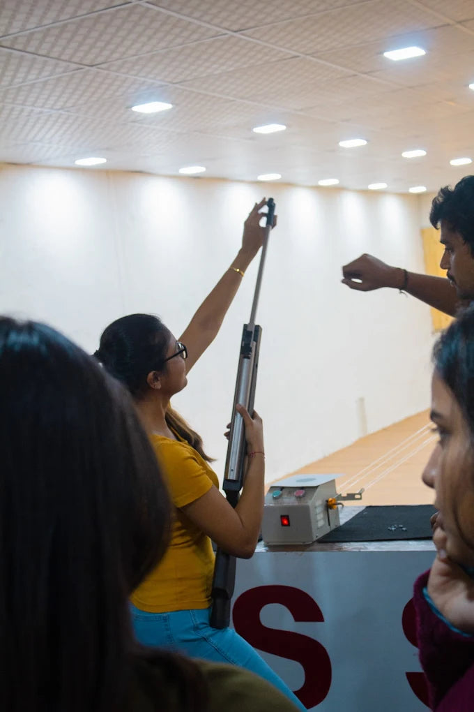 A young woman practices with a rifle at a shooting range, observed by others.