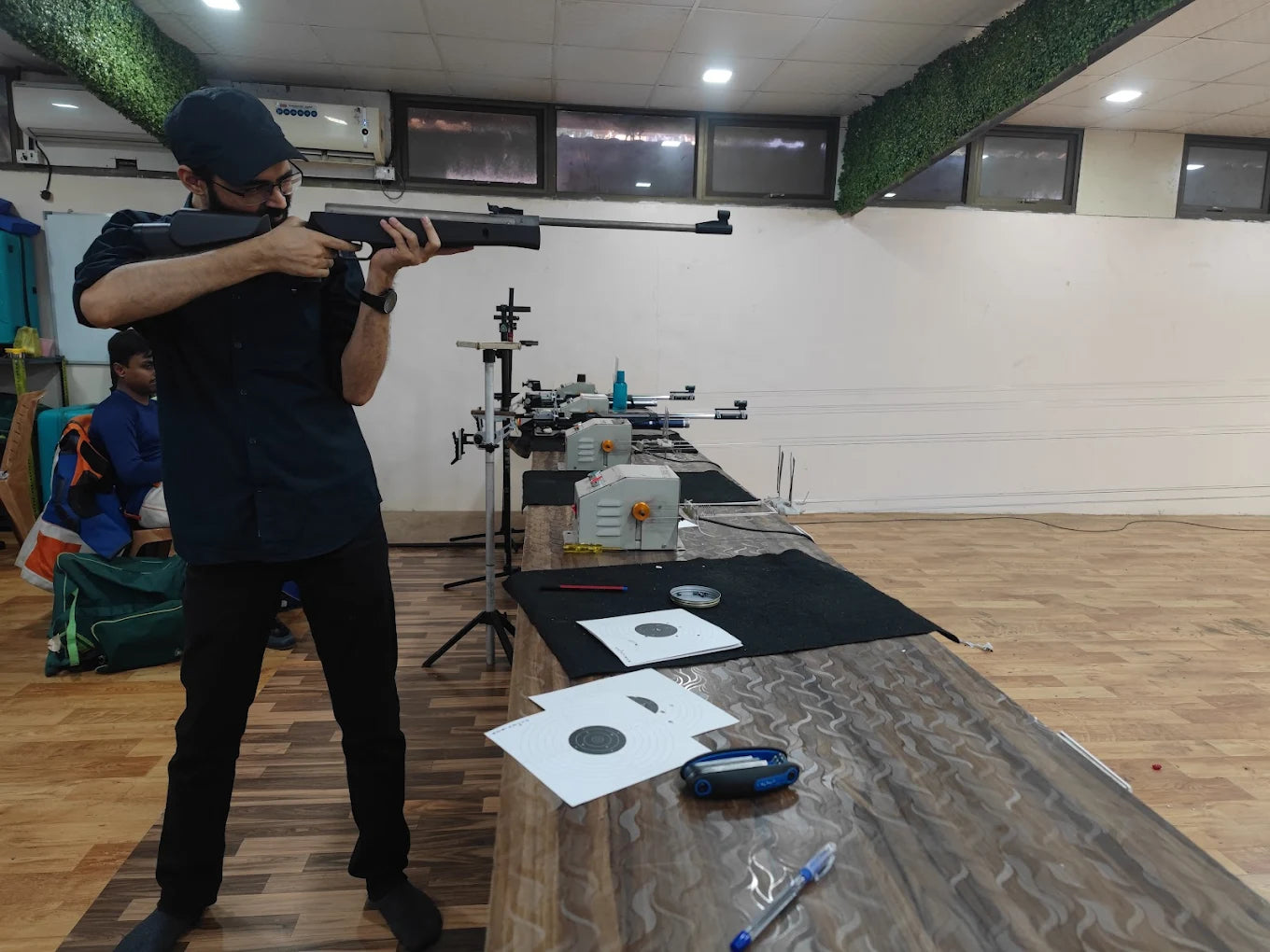 A young man aims a rifle at a shooting range, with targets and equipment visible on the table.