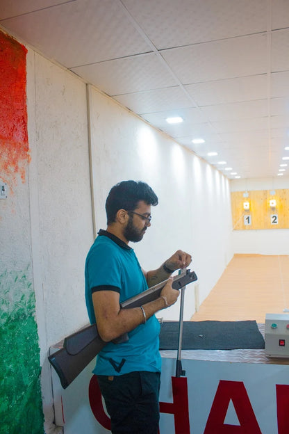 A man in a blue shirt prepares a rifle at a shooting range. Targets are visible in the background.
