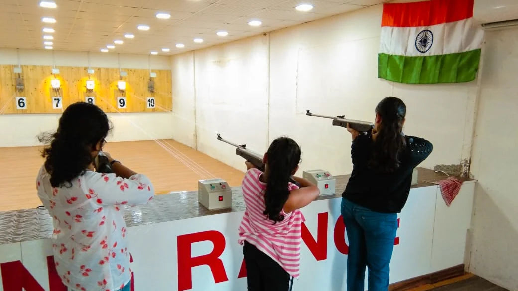 Three young women practice shooting at a target range, with an Indian flag hanging in the background.