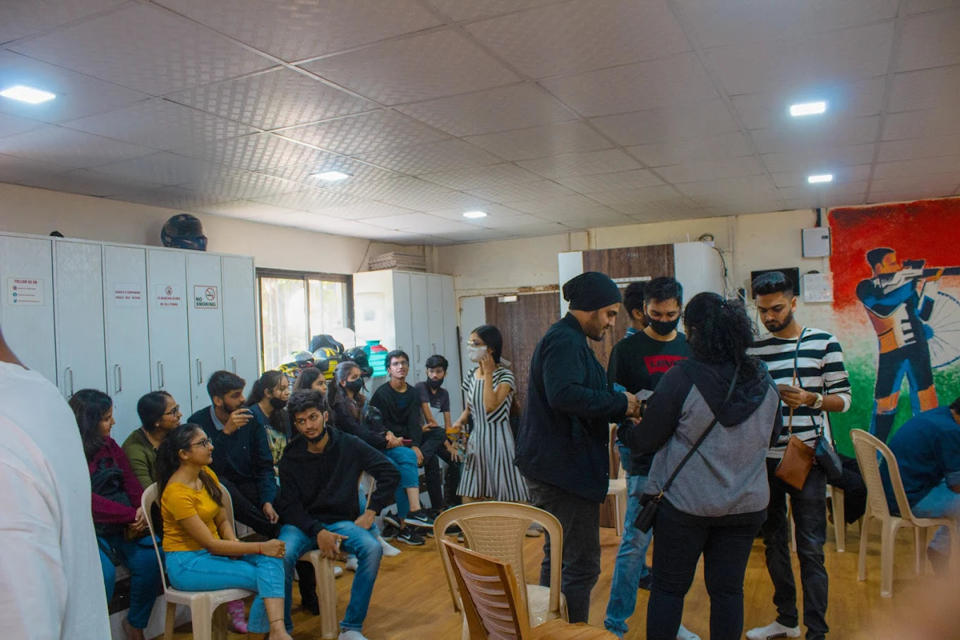 A group of young adults gather in a room with lockers and a mural, some standing and interacting, others seated.