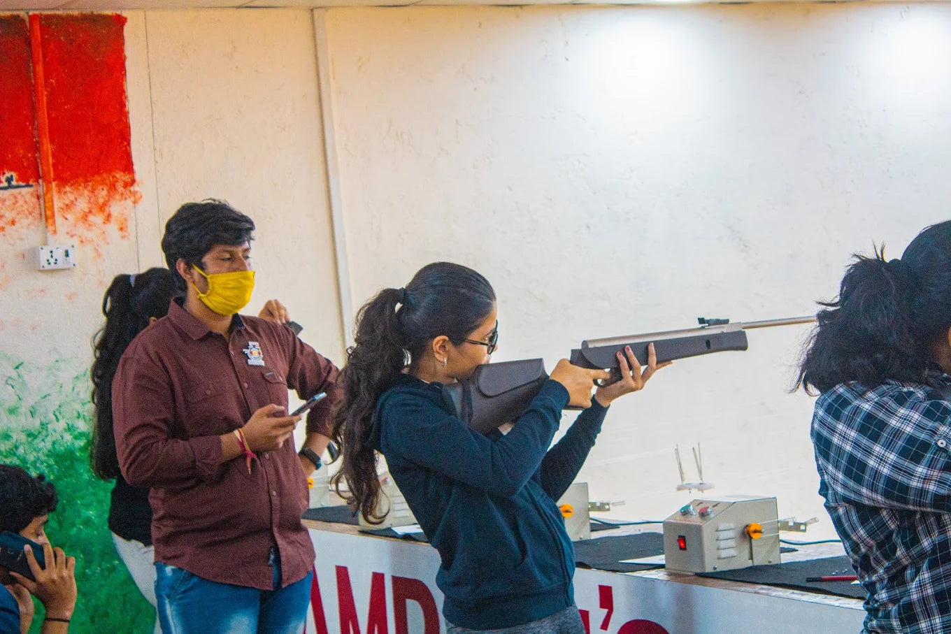 A young woman aims a rifle at a shooting range while a young man in a mask watches.