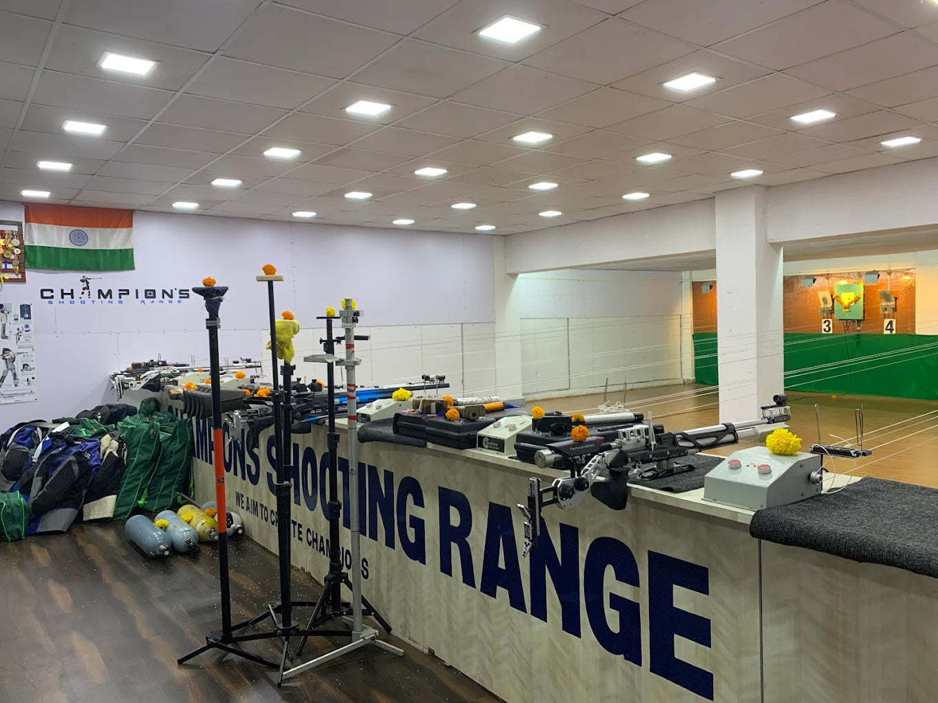 Indoor shooting range with rifles on a counter, targets in the background, and an Indian flag.