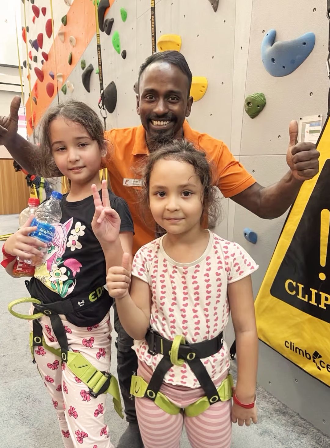 Two young girls and an adult pose in front of a climbing wall, both girls wearing harnesses and giving thumbs up.