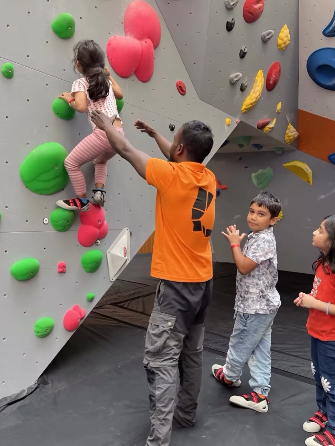 A man helps a young girl climb a rock wall while two other children watch.
