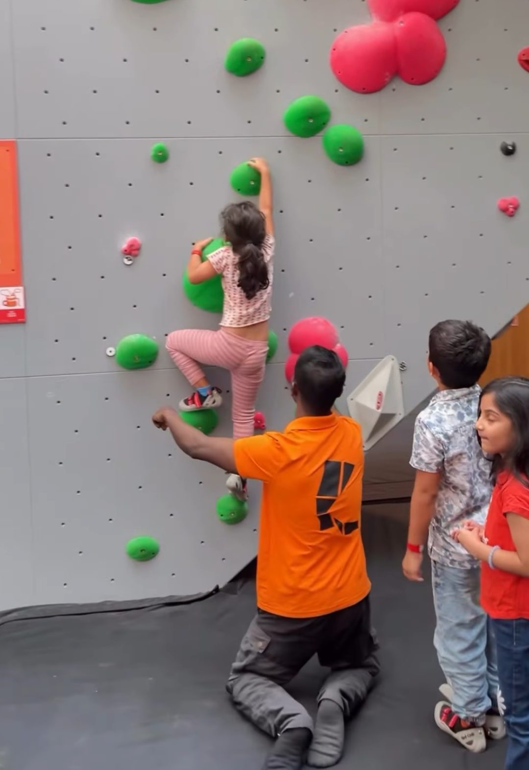 A young girl climbs a bouldering wall with an instructor and other children watching.