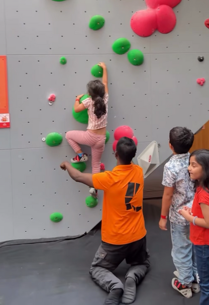 A young girl climbs a bouldering wall with an instructor and other children watching.