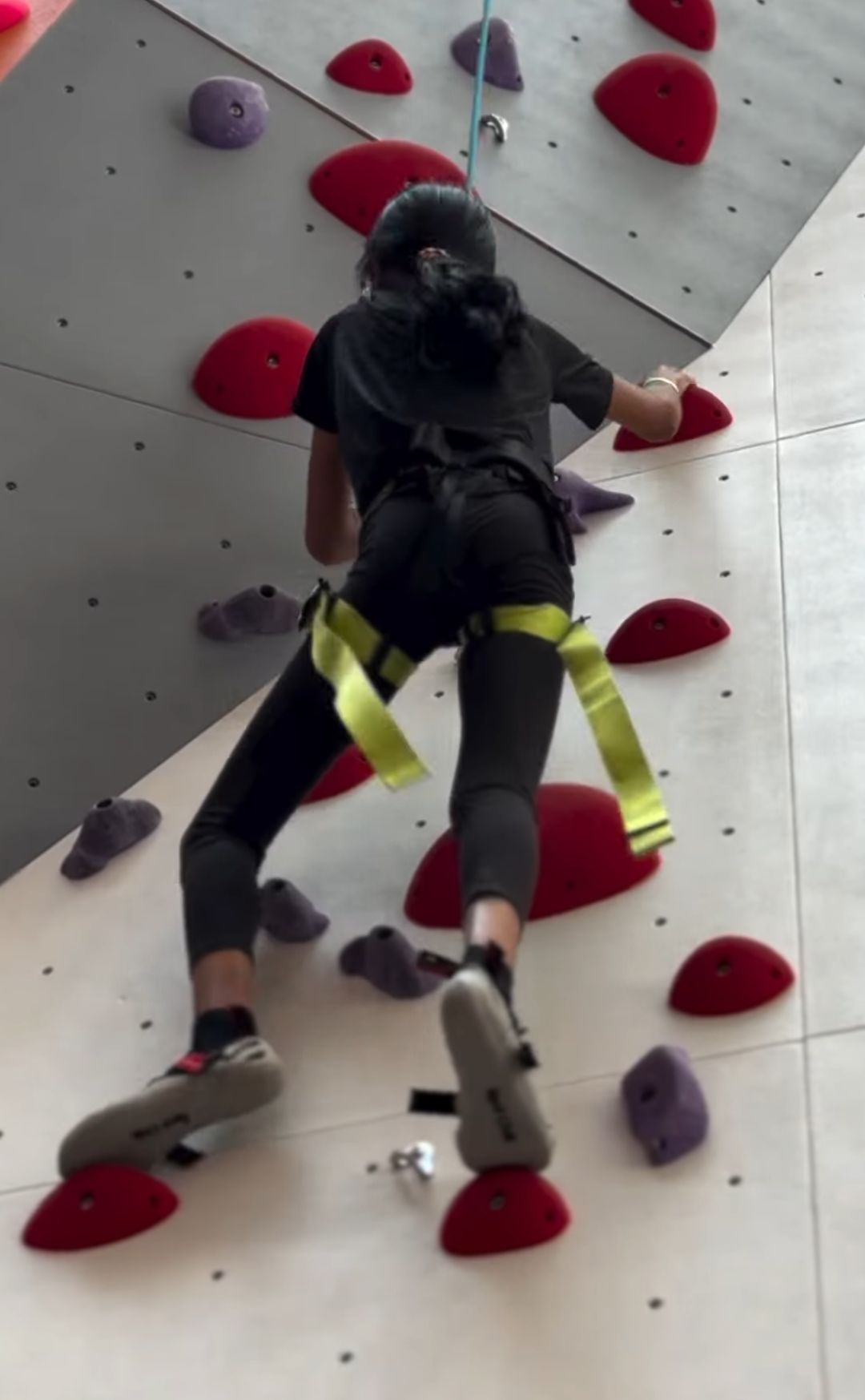 A young person climbs a colorful indoor rock wall, wearing a harness and climbing shoes.