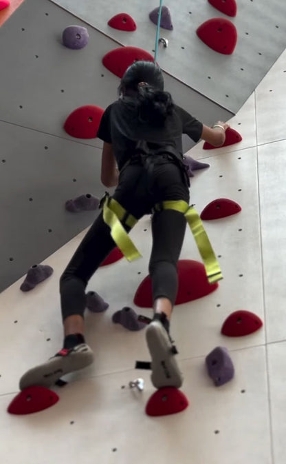 A young person climbs a colorful indoor rock wall, wearing a harness and climbing shoes.