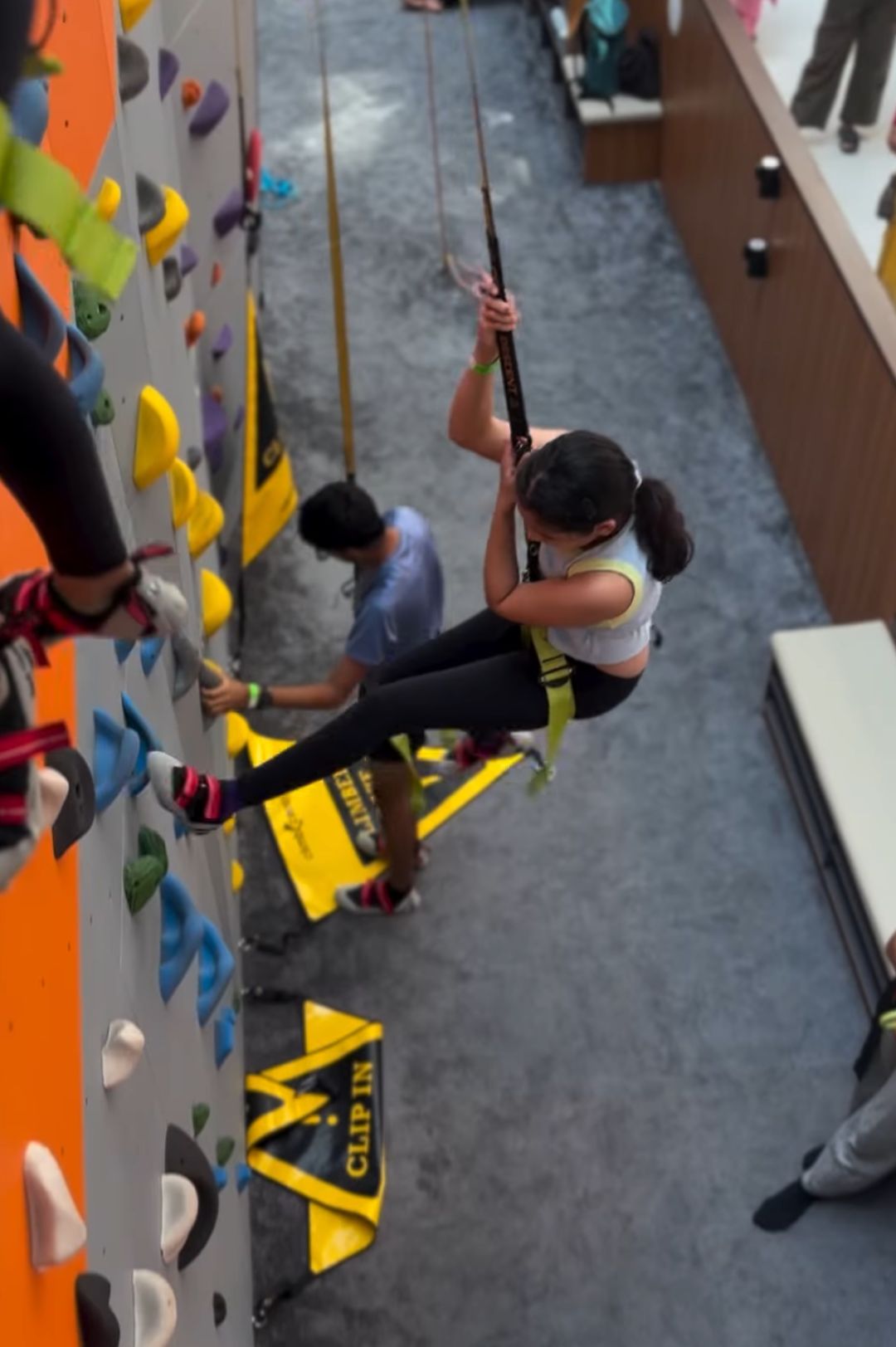 A young girl is belayed while climbing an indoor rock wall, with another person on the wall below her.