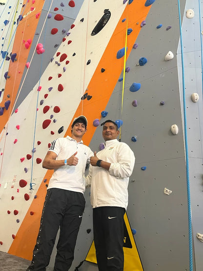 Two adult men pose in front of a colorful indoor climbing wall.