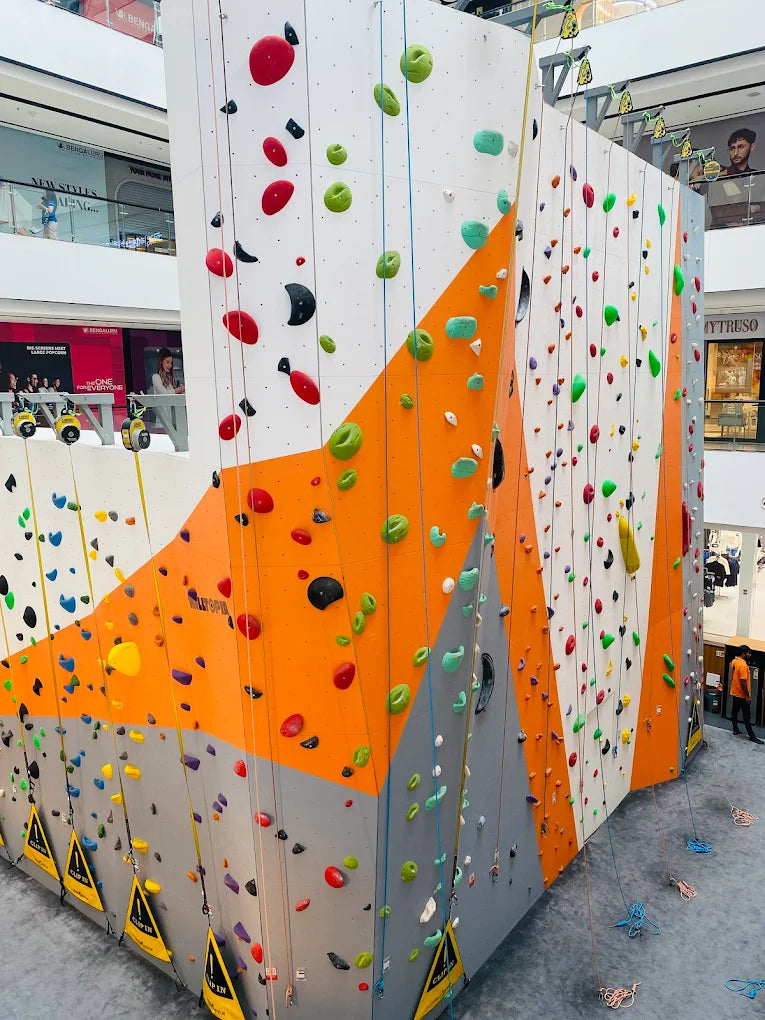 A colorful indoor climbing wall with ropes and holds, and a few people in the background.