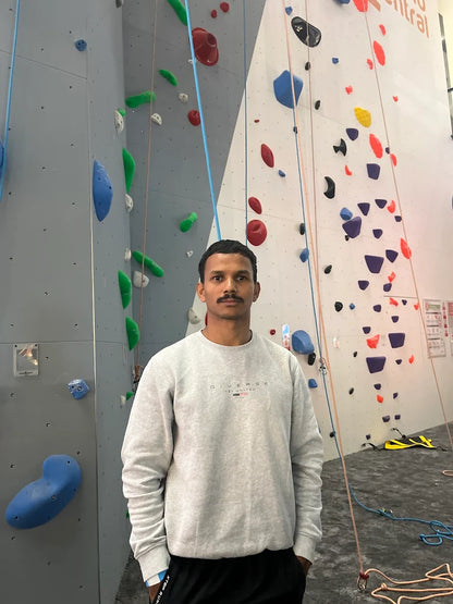 A young man stands in front of a climbing wall with colorful holds.