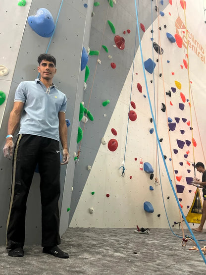 A young man stands at the base of a climbing wall in an indoor gym. He is wearing athletic clothing and climbing shoes.