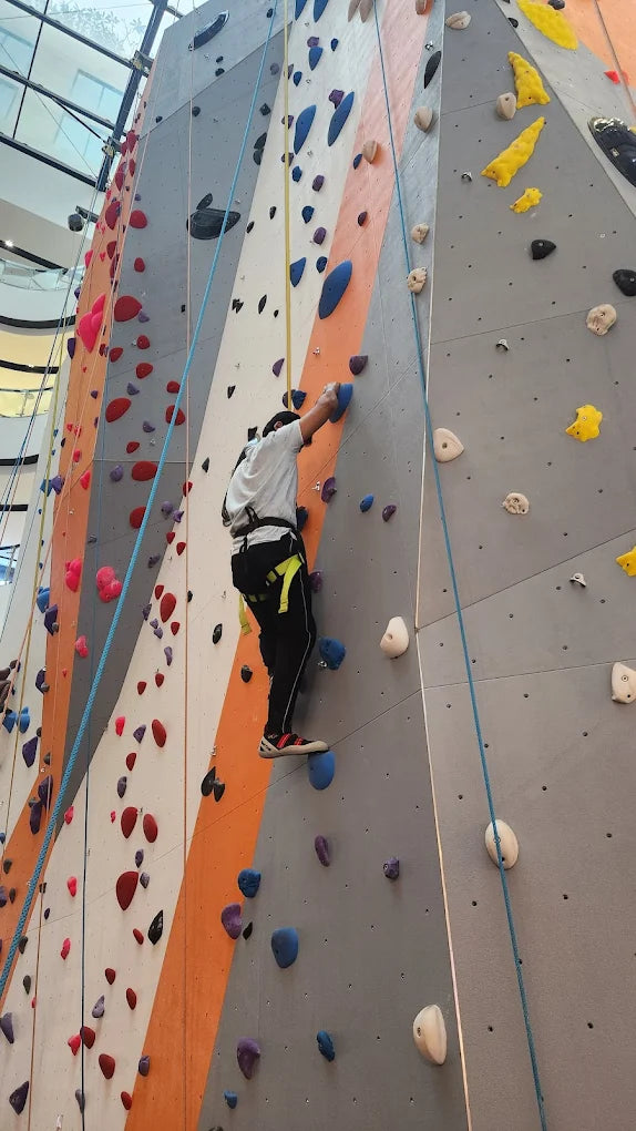 A young person climbs a colorful indoor rock wall, using ropes and colorful holds.