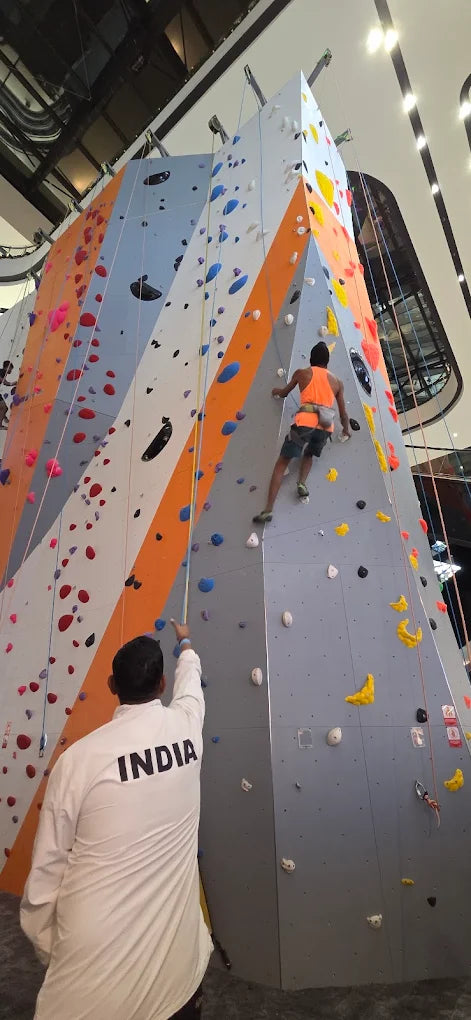 A young man climbs a colorful indoor rock wall while another person in a white jacket with "INDIA" on it belays him.
