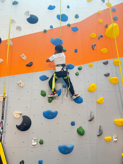 A young child climbs an indoor rock wall, wearing a harness and helmet.