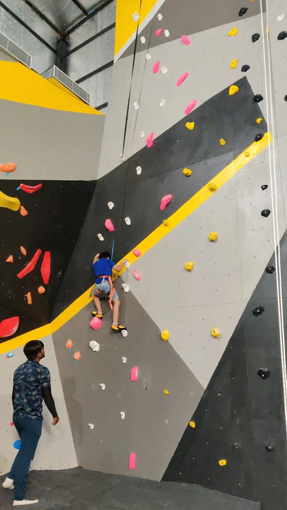 Child wearing safety harness climbing tall indoor rock wall with colorful holds while instructor watches below