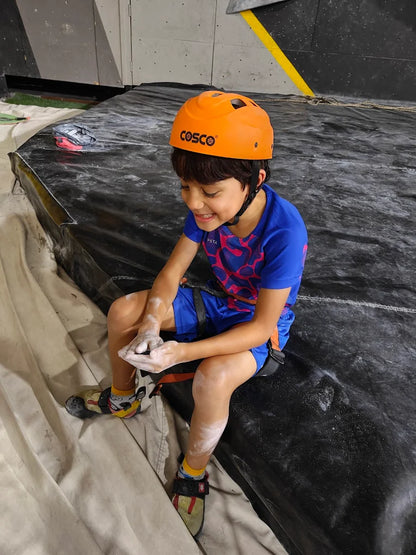 Child wearing a Cosco helmet sitting on a black mat, preparing to climb.