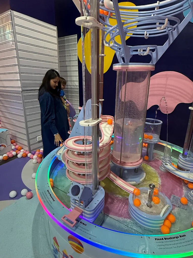 A woman holds a child while watching a colorful water play exhibit with orange balls.