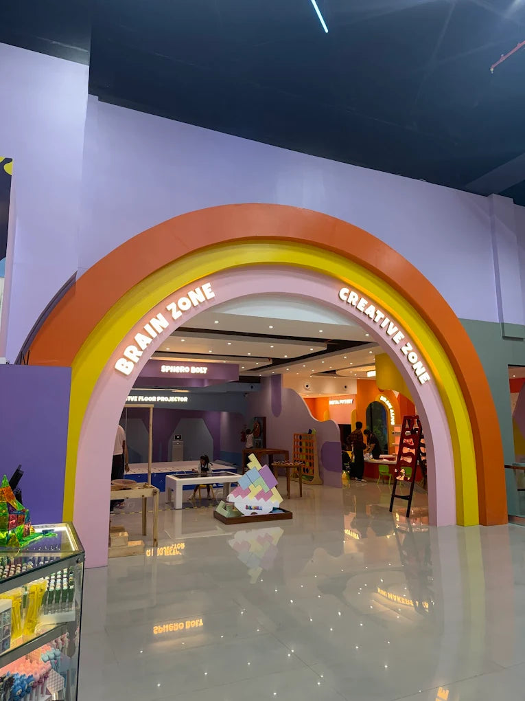 Children play in a colorful indoor play area with a rainbow archway labeled "Brain Zone" and "Creative Zone."
